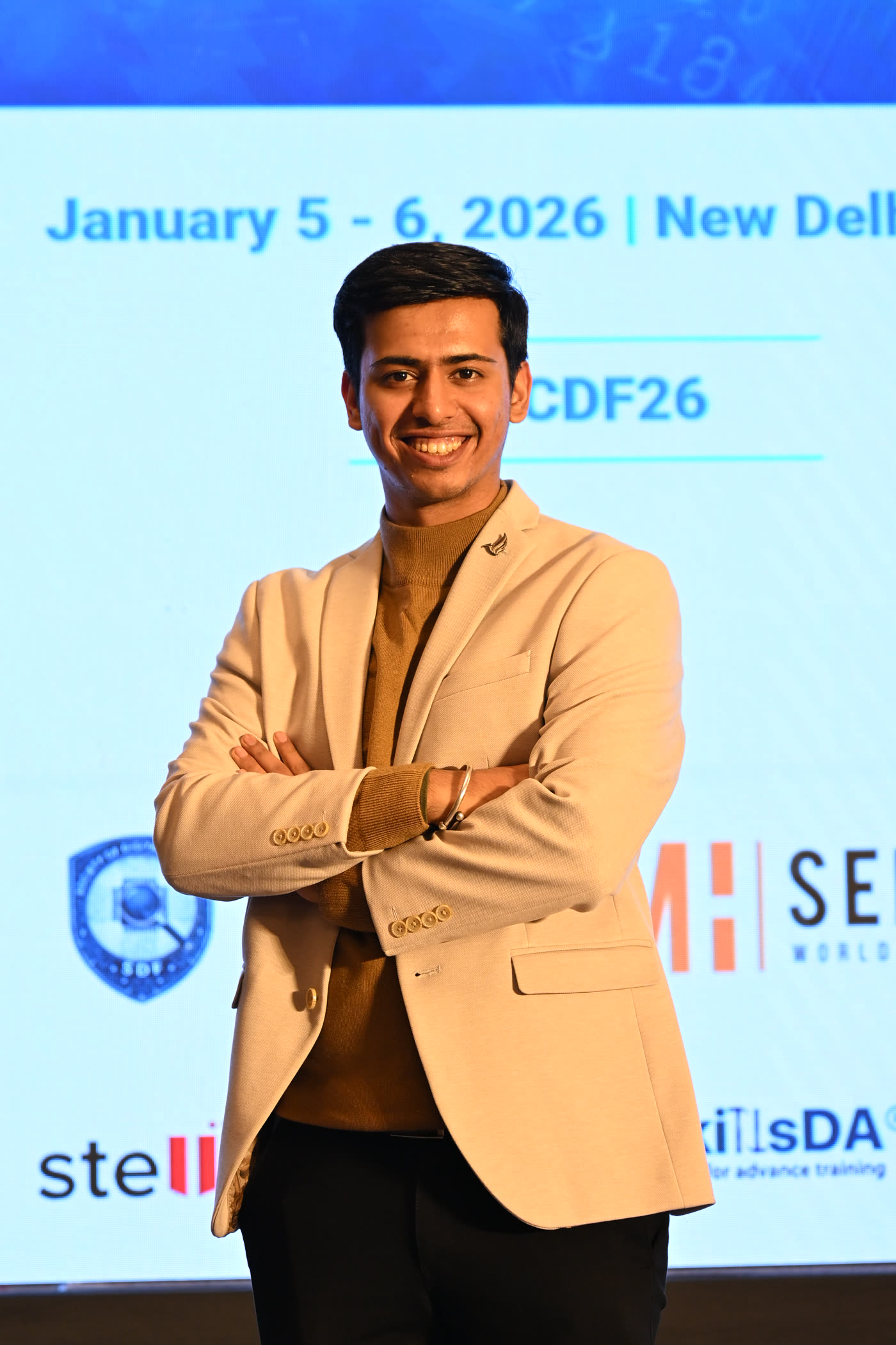 Aryan in a beige blazer, arms crossed, on stage at a security event in New Delhi