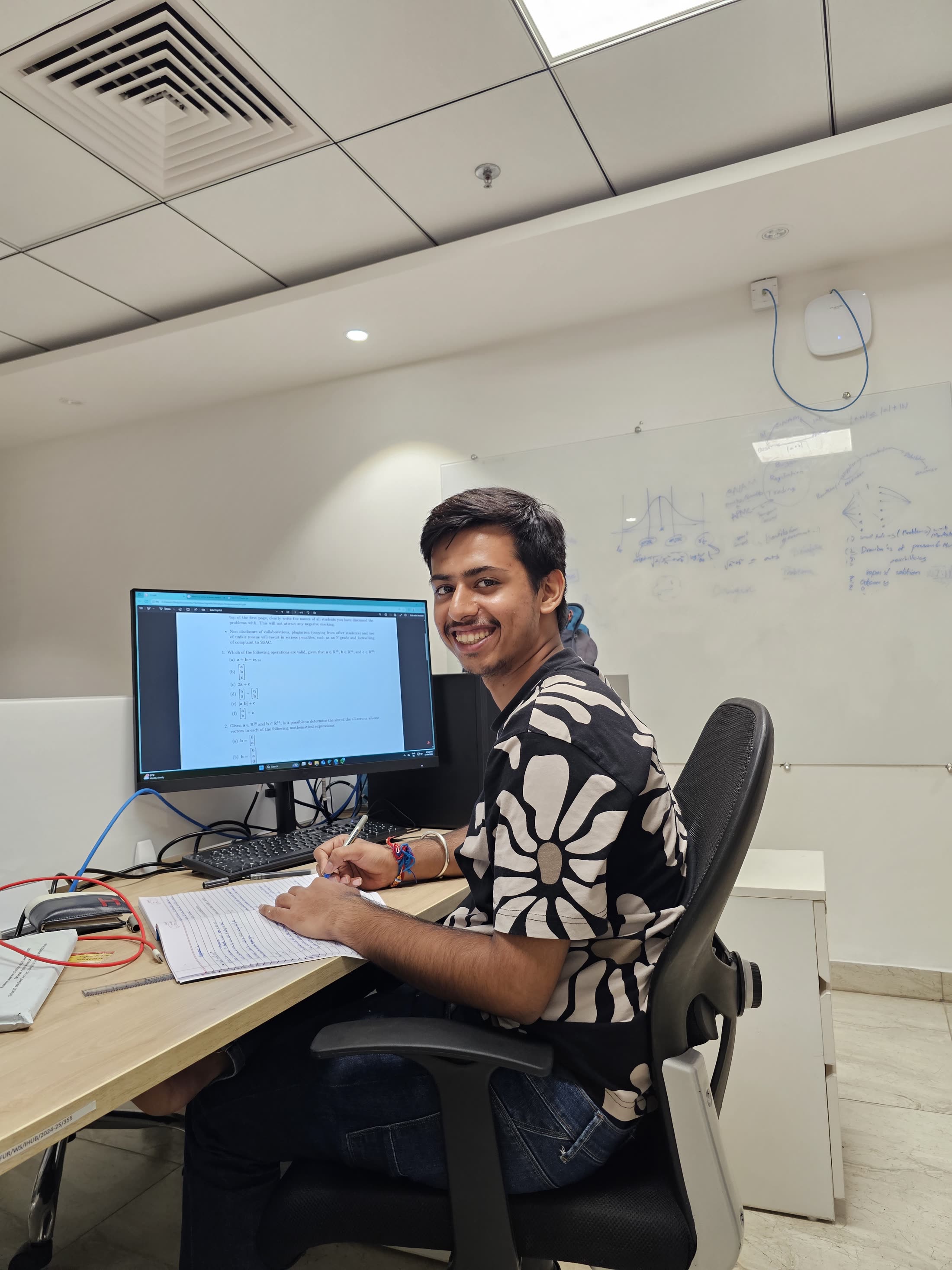Aryan at his desk in the lab, turned towards the camera, a whiteboard of notes behind him